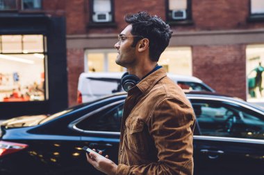 Gün ışığında şehir caddesinde yürürken kulaklık ve gözlük takan yakışıklı, sıradan yetişkin bir adamın yan görüntüsü.