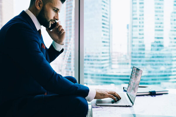 Side view of crop manager in dark suit browsing laptop and talking on smartphone while sitting near table in contemporary office