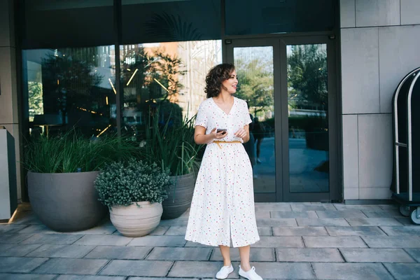 Αισιόδοξη Γυναίκα Κομψό Λευκό Φόρεμα Κρατώντας Smartphone Στο Χέρι Ενώ — Φωτογραφία Αρχείου