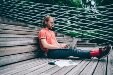 Şehir parkında bacakları açık, tahta bir kürsüde otururken dizüstü bilgisayarda çalışan genç bir adam. 