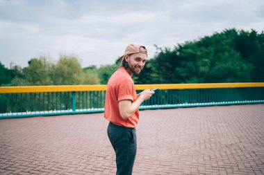 Neşeli, güler yüzlü, sıradan giyinmiş, şapkalı, sosyal ağda dolaşmak için cep telefonu kullanan sokakta kameraya bakan yakışıklı adam.