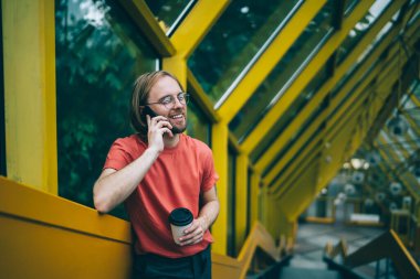 Camdan duvarın yanında duran kıllı genç adam, kapalı yaya köprüsünde elinde kahve fincanı tutarken telefonla konuşuyor ve parmaklıklarla konuşuyor.