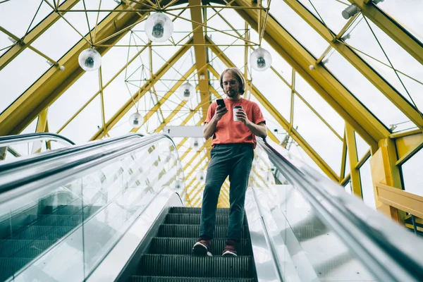 カジュアルな服装の男性と 街を歩くエスカレーターの上で友人とチャットするために携帯電話を使用してコーヒーを飲むメガネに焦点を当てた — ストック写真