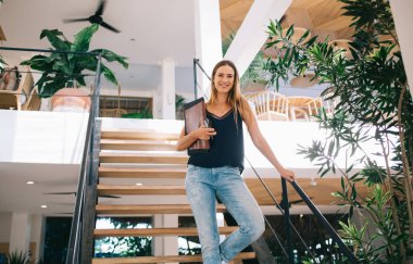 Gündelik modaya uygun giyinmiş, gülümseyen beyaz bayan öğrencinin yarım boy portresi modern iç kampüste aşağı katta yürüyor, 20 'li yaşlarda, gülümseyen genç iş kadını kameraya bakıyor.