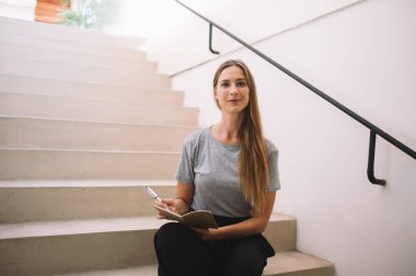 Not defterinde kendinden emin beyaz kadın yazısının yarım boy portresi. Yirmili yaşlarda kampüsteki merdivenlerde oturan yeniyetme öğrenci öğrenme ve yazma konusunda ciddi fikirlere dikkat çekiyor.