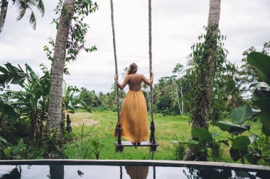 Turuncu yaz elbisesi giymiş, tropik yemyeşil bahçede salıncakta sallanan, bulutlu sahilde tatil yapan bir kadının arka görüntüsü. 