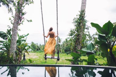 Turuncu yaz kıyafetli modern bir kadın plaja karşı yeşil çimlerle tropikal bahçede sallanıyor ve tatilin tadını çıkarıyor. 