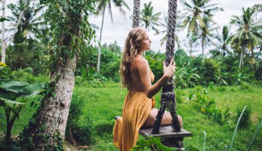 Portakal rengi yaz kıyafetli modern sarışın bayanın çimenli palmiye bahçesinde salıncakta sallanıp tatilin tadını çıkarışının görüntüsü.