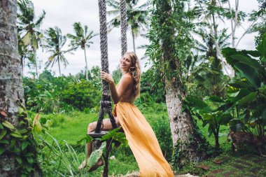 Yeşil palmiye bahçesinde salıncakta sallanan turuncu yazlık elbiseli, gülümseyen sarışın, kıvırcık kadın. 