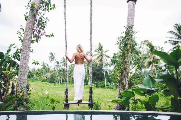 Beyaz yaz elbisesi giymiş, tropikal bahçede salıncakta sallanan ve tatilin tadını çıkaran kıvırcık, modern bir kadın. 
