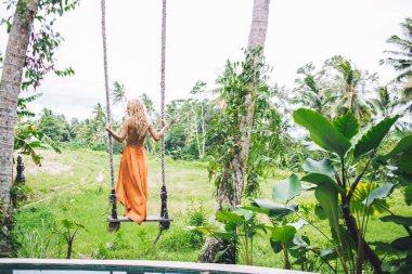 Turuncu yaz kıyafetli, tropikal korulukta salıncakta sallanan, tatilin tadını çıkaran, kıpır kıpır, genç, modern bir kadının manzarası.