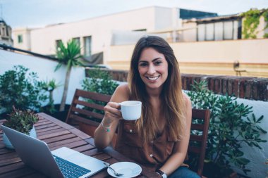 Kafede dizüstü bilgisayar başında oturan ve kameraya neşeyle bakarken sıcak içecek içen rahat bir kadın.