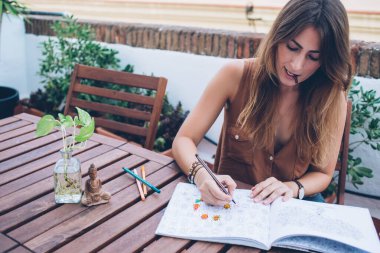 Genç odaklı kadın boyama çizimleri stres önleme kitabında ve terasta masada otururken keçeli kalemin kepini dişlerinde tutarak.