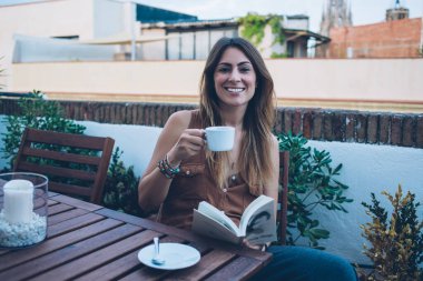 Sıcak içecekten zevk alırken ve açık kafede güzel kitaplar okurken günlük kıyafetlerle gülümseyen ve kameraya bakan pozitif genç bir kadın.