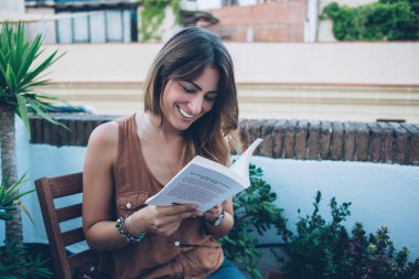 Çekici, mutlu, günlük kıyafetlerle kitap okuyan bir kadın. Terasta, yeşil çiçek ve ağaçların arasında, bulanık kentsel çevreye karşı otururken zevkle kitap okuyor.