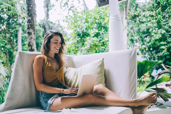 美しい幸せなブルネット女性でカジュアル服作業ノートパソコン上で座っている間白いソファに庭で晴れた日 — ストック写真