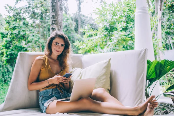 携帯電話でメッセージを楽しみ 庭のラップトップで白いソファに座っている間カメラを見てカジュアルな服の美しい幸せなブルネットの女性 — ストック写真