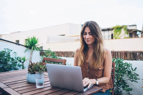 Cheerful woman with wristlet watch browsing laptop sitting on wooden chair on terrace while looking at screen in thoughts and working