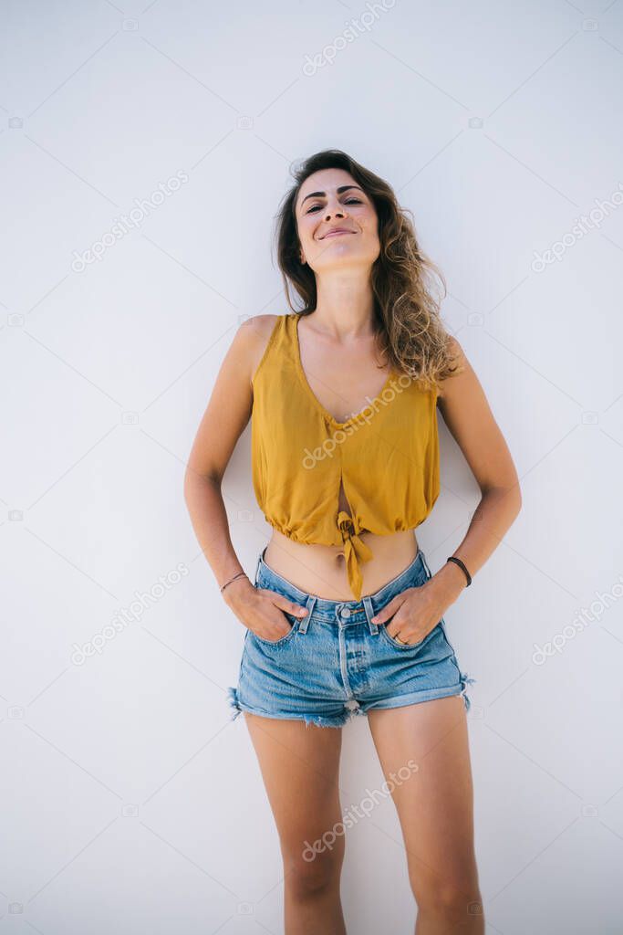 Pleased relaxed carefree casual young trendy woman in top and shorts leaning on white wall on street on sunny day