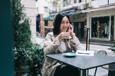 Kahve fincanı ve kısa saçlı genç Asyalı kadın masada oturmuş kameraya bakarken akıllı telefonla konuşuyor ve şehir yoluna gülümsüyor.
