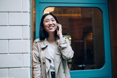 Neşeli gülümseyen, modaya uygun, trençkotlu genç Asyalı kadın telefonda konuşurken gündüz vakti kapı eşiğinde dikiliyor.