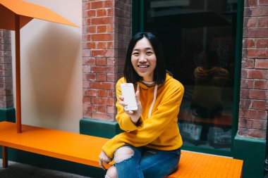 Sarı kapüşonlu genç, neşeli etnik kadın turuncu bankta oturuyor kameraya gülümsüyor ve boş beyaz ekranlı akıllı telefonu gösteriyor.