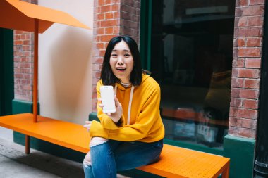 Sarı kapüşonlu ve yırtık pantolonlu genç, şaşkın, kısa saçlı Asyalı kadın kameraya telefon ekranını gösteriyor.