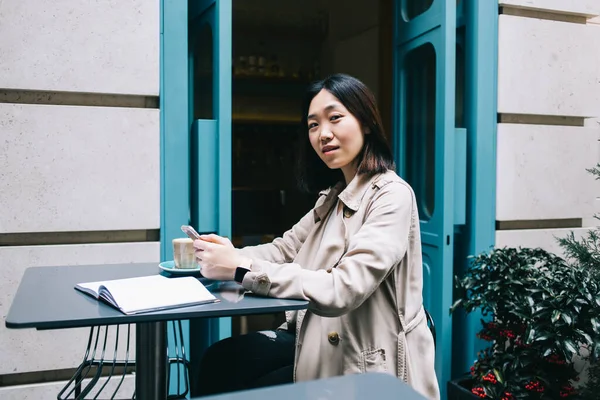 一个略带亚洲笑容的黑发年轻女子坐在桌旁 拿着笔记本 用智能手机在街上的咖啡店里看着相机 — 图库照片