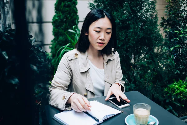 以认真的亚裔女学生为对象 穿着夹克 带着笔记本在智能手机上浏览 并在咖啡店的阳台上享受饮料 — 图库照片