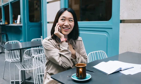 Young cheerful woman speaking on cellphone and smiling while sitting in open air cafe with served coffee and notebook on daytime