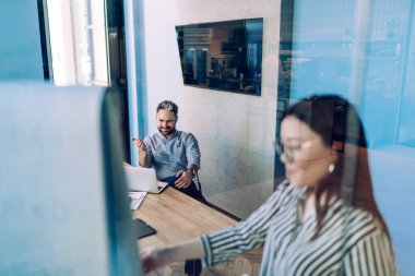 Erkek, modern şirket ofisinde toplanma projesini tartışırken gülümseyen ve meslektaşını dinleyen bir dizüstü bilgisayarla masada oturuyor.