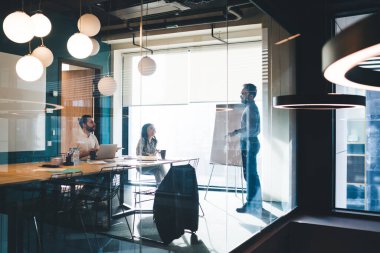 Laptoplu sakallı bir adam, siyah saçlı bir kadınla masada oturuyor ve pencerede duran gömlek ve pantolonlu patronu dinliyor.