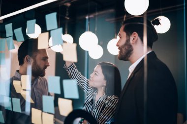 Siyah saçlı sakallı adamlar çizgili siyah beyaz tişörtlü Asyalı kadını dinliyorlar. Cam duvardaki renkli etiketi gösteriyorlar.