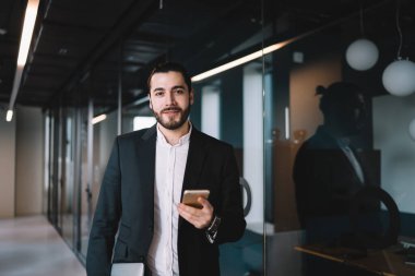 Genç, kendine güvenen, siyah takım elbiseli, beyaz gömlekli, akıllı telefonu elinde tutan ve ofis koridorunda dikilirken kameraya bakan erkek yönetici.