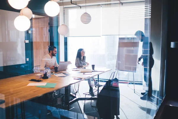 Businessman standing next to window and presenting idea to colleagues who sitting at table with laptop and office supplies in contemporary workplace