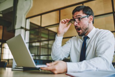 Optik gözlüklü beyaz erkek girişimci, modern laptop cihazından haber almaktan şoke oldu. Gözlüklü 30 'lu yaşlarda şaşkın işadamı, ofis içi web tarayıcıları için çalışma saatlerini merak ediyor.