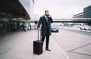 Tüm vücudu, takım elbiseli, bavullu, akıllı telefondan konuşan ve havaalanı terminalinin yanına bakan bir erkek.