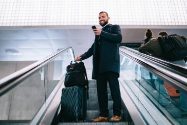 Havalimanında cep telefonuyla sohbet ederken bavuluyla yürüyen merdivende duran siyah ceketli mutlu bir erkek.