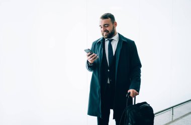 Takım elbiseli, akıllı telefon kullanan, gülümseyen ve beyaz arka planda duran havaalanı duvarına bakan mutlu genç adam.