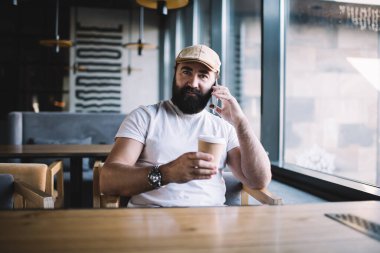 Ciddi sakallı, modaya uygun şapkalı, kol saati ve bir fincan sıcak içecekle şık giyinmiş akıllı telefondan konuşan ve kameraya bakan biri.