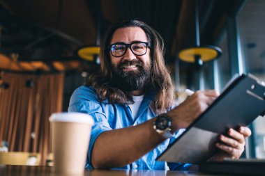 Orta yaşlı, kol saati takan, gözlüklü, kafede rahat bir masada otururken fikirlerini not eden mutlu bir adamdan.