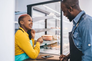Koyu renk önlüklü Afro-Amerikan genç bir adamın pastayı seçmesi ve sarı gömlekli ve turkuaz önlüklü şık siyahi bir kadınla konuşması. 