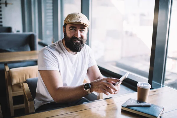 Счастливый Бородатый Мужчина Кепке Помощью Смартфона Сидя Современном Кафе Кофе — стоковое фото