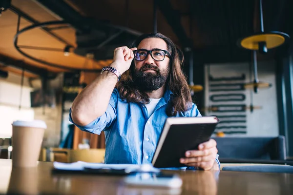 Handsome mature man in eyewear for vision correction holding interesting book spending free time on hobby, smiling prosperous business owner in casual wear sitting in cafe with notebook planner