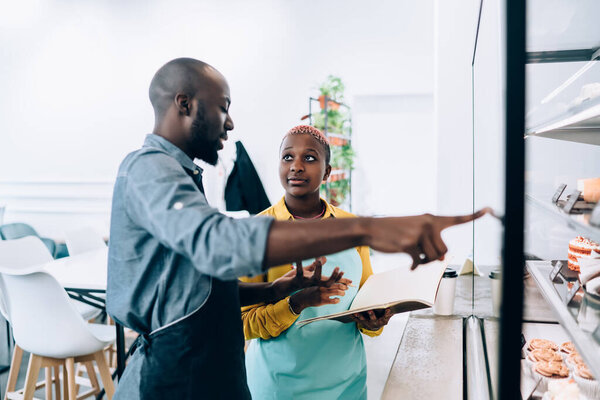 African American young modern smiling cheerful colleagues with notebook chatting with each other and pointing to counter with desserts in cafeteria in city