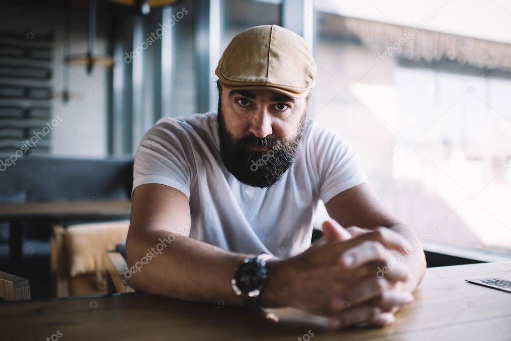 Portrait of handsome bearded male in hat and casual wear sitting at table in cafe interior, confident determined 40 years man looking at camera spending time in coffee shop owning small business
