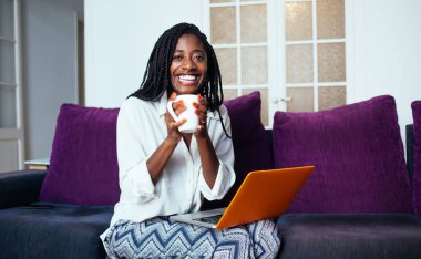 Kahkahalarla gülen, elinde çay fincanı tutan neşeli, esmer bir kadın kanepenin içinde rahat bir şekilde çalışmaktan zevk alan, evde dinlenen, kahve içen ve dizüstü bilgisayar kullanan genç ve zengin bir kadın.