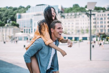 Güneşli bir yaz gününde şehir meydanında yürürken birlikte eğlenen ve mutlu anların tadını çıkaran neşeli, gülen genç çiftin yan görüntüsü.
