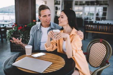 Kafe masasında birlikte oturan mutlu çift ellerinde içecek dizüstü bilgisayarı ve boş ekranlı akıllı telefonuyla kız arkadaşını dikkatlice dinlerken ve açıklarken el kol hareketi yaparak kız arkadaşına sarılıyor.