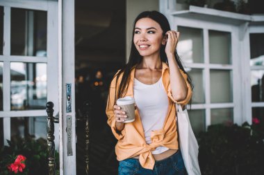 Neşeli, şık, esmer, etnik bir kadın bir fincan kahveyle kafenin dışına çıkıyor, saçlarına dokunuyor ve gündüz gözlerini kaçırıyor.